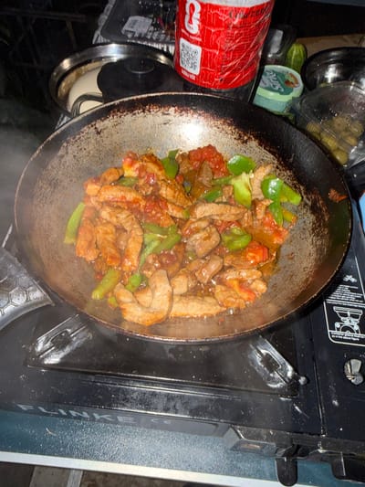 A close-up, overhead shot captures stir-fried meat and vegetables cooking in a wok. The dish features strips of browned meat, green bell pepper slices, and red tomato chunks, all coated in a reddish-brown sauce. The wok itself is seasoned and appears to be well-used. It rests on a black portable camping stove. In the background, a red beverage container with visible damage and a QR code is present, along with a small green cucumber and containers of food. The overall setting suggests an outdoor cooking scenario, potentially in the city of Kanjoši, Montenegro, given the context. The lighting is dim, contributing to a focus on the food and cooking equipment.