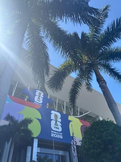 The image shows a vibrant mural for the FIFA World Cup 2026 adorning the exterior of a stadium in Miami Gardens, Florida, United States of America. Two palm trees, one on the left and another on the right, partially obscure the view of the building, their fronds reaching towards a clear blue sky. The mural features bold, colorful graphics, including a large blue section with the FIFA World Cup trophy and the text "FIFA WORLD CUP 2026." The stadium itself has a modern design with a white, angled roof structure. An American flag is visible flying on a flagpole in front of the mural. The scene is bathed in bright sunlight, casting some lens flare into the image.