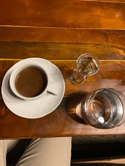 An overhead view captures a brown planked wooden table surface in Dulovine, Montenegro. On the left, a white ceramic cup containing dark brown liquid rests on a matching white saucer. To the right of the saucer, a clear stemmed glass is positioned, containing a clear liquid. Below and to the right of the stemmed glass is a larger, clear, cylindrical glass, also containing clear liquid, with a small white object visible at its base. The bottom left quadrant of the image shows a portion of a human leg clad in light-colored trousers, indicating a person is seated at the table. The lighting creates reflections on the glasses and highlights the wood grain.