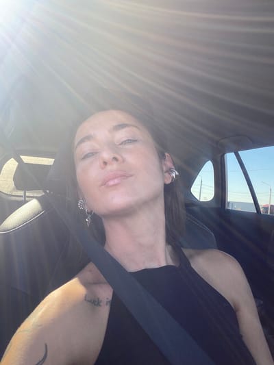 The image displays a female individual seated within a vehicle, secured by a black seatbelt, with the setting identified as the city of Sheragul, Russia. She has fair skin, light-colored eyes, and brown hair, looking directly at the camera with a neutral expression and slightly parted lips. Her attire consists of a black sleeveless top. She wears multiple silver-toned earrings: several hoops/cuffs on her left ear and two distinct pieces on her right ear, one appearing spherical and the other a dangling element with a pointed, conical shape. A black script tattoo, partially legible as "luck is," is visible on her left upper arm, accompanied by a crescent moon-like design. Bright sunlight enters the vehicle from the upper left, causing lens flare and strong illumination. Through the vehicle's side and rear windows, a clear blue sky, distant structures, and power lines are visible, indicating an outdoor, possibly urban or semi-urban, environment during daylight hours.