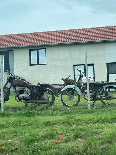 The image depicts a scene in an outdoor setting. Three motorcycles, appearing to be in a state of disrepair, are visible behind a chain-link fence. Behind the fence, a building with a tiled roof and several windows is present. The surrounding environment consists of green grass. The image was presumably taken in Ushintsi, Bulgaria. There are no people present in the image.