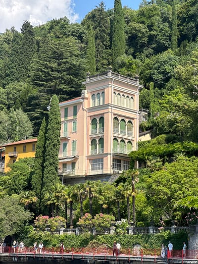 This image depicts a picturesque scene in Varenna, Italy, with a focus on a charming, light pink villa nestled amongst lush green foliage. The villa features multiple stories with arched windows, many of which are adorned with green shutters. The architectural style suggests a European influence, possibly Italian.
The scene is bathed in natural light, indicating it was taken during the daytime, likely late morning or afternoon, given the shadows. The weather appears to be pleasant, with a clear blue sky visible in the upper left portion of the image.
In the foreground, a red railing lines a walkway or balcony overlooking a body of water, though the water itself is not clearly visible. Several people are casually strolling along this area, some dressed in light-colored clothing, enjoying the scenic view. One individual stands out in a light blue top on the right side of the image.
The surrounding environment is abundant with diverse plant life. Tall, slender cypress trees stand prominently on the left, while palm trees and flowering bushes add to the tropical yet Mediterranean ambiance. The dense greenery creates a sense of seclusion and natural beauty around the villa.
To the lower right, a glimpse of another building, a small yellow structure with windows, suggests an urban or village setting integrated with nature. The overall mood of the image is serene and inviting, capturing a tranquil moment in a beautiful Italian location. There is no visible text in the image.