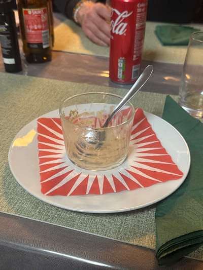 The image depicts a dining table located in Lalden, Switzerland. In the foreground, a clear glass bowl containing light brown food residue and a silver spoon is centered on a white ceramic plate. A folded orange and white patterned paper napkin is placed under the glass on the plate. The table surface is covered by a textured light green material, which partially overlays a darker, reflective grey surface. In the blurred background, from left to right, a dark bottle, a brown Sagres beer bottle, a red Coca-Cola 330ml can, and an empty clear drinking glass are visible. A folded dark green napkin is situated to the right of the Coca-Cola can. A human hand, adorned with a multi-colored beaded bracelet, is partially visible behind the Coca-Cola can, indicating an individual is seated at the table. The scene suggests recent consumption of a meal or dessert.