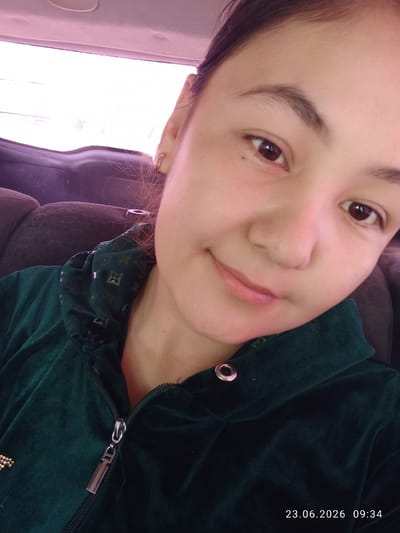 A close-up, selfie-style photograph shows a young woman with brown eyes and dark hair pulled back from her face. She is wearing a dark green velour hooded jacket with a zipper down the front and decorative elements on the left chest. The background suggests she is inside a vehicle, with a dark-colored seat visible and the interior ceiling of the car, which has a light-colored, illuminated lining. The image is timestamped "23.06.2026 09:34". The context of Batken City, Kyrgyzstan, is not visually apparent from the image itself.