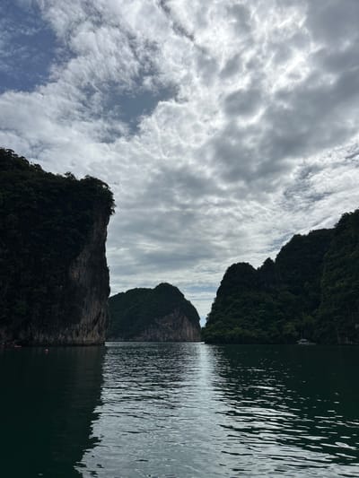 The image depicts a tranquil scene in Khlong Khian, Thailand, characterized by lush green karst islands rising from dark, rippling water under a cloudy sky. The foreground is dominated by the water's surface, reflecting the overcast sky and the imposing silhouettes of the islands. On the left, a sheer, dark cliff face covered in dense vegetation drops dramatically into the sea. In the distance, the water opens to reveal two more rounded, tree-covered islands. The sky is filled with a mix of white and gray clouds, with patches of blue peeking through, suggesting a dynamic weather pattern. A white sailboat is visible near the island on the right. The overall atmosphere is one of natural beauty and serenity, with a hint of the majestic geological formations that define the region.