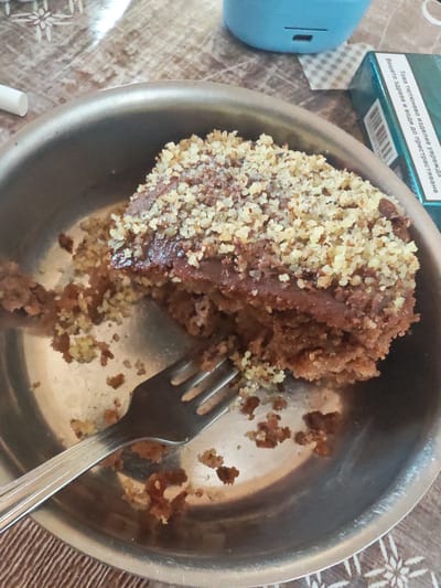 The image shows a slice of cake in a metal bowl. The cake has a chocolate frosting layer topped with chopped nuts. A fork is partially embedded in the cake, indicating someone has been eating it. There are crumbs scattered around the cake and on the bottom of the bowl. A pack of cigarettes and a blue device sit to the right of the bowl. The table underneath has a pattern on it. While the image was provided with the information that the location is in the city of Bogdanovo, Bulgaria, this cannot be forensically determined from the image itself.
