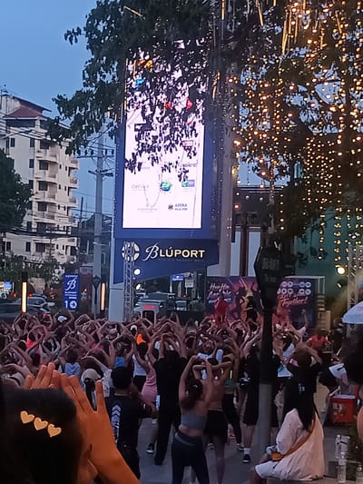 A crowd of people are gathered for an outdoor aerobic exercise session in Hua Hin City Municipality, Thailand. Many of the participants are holding their hands above their heads in a heart-shaped gesture. A large electronic screen displays an image and text, and is positioned in front of a building identified as "Blúport." The scene is illuminated by strings of lights hanging from trees. A smaller sign is visible, with a "B" logo and the number "10." The setting suggests a public space or plaza in an urban area. Some buildings are visible in the background.