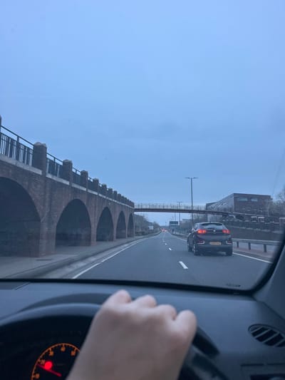 The scene depicts a roadway from the perspective of a vehicle's driver, looking eastward on Pier Road in Gillingham, Kent, United Kingdom. The observer's right hand grips the steering wheel of a right-hand drive vehicle, with a partially visible orange-lit dashboard dial showing a red indicator.
The environment is an urban road under an overcast, light blue-grey sky. A two-lane asphalt carriageway extends into the distance, marked with white dashed lane lines. To the left, a multi-arch brick railway viaduct runs parallel to the road, featuring a dark metal railing atop a brick parapet with decorative stone finials. The underside of the arches is dark. To the right, a dark-colored SUV (license plate GM18 FXN) travels in the right lane, its rear lights illuminated. Beyond this vehicle, a reddish-brown pedestrian bridge with metal railings spans across the road. Further right, a multi-story building with dark brickwork and windows is visible, likely part of a commercial or industrial complex. Multiple streetlights are present along the roadside. No other individuals are visible.