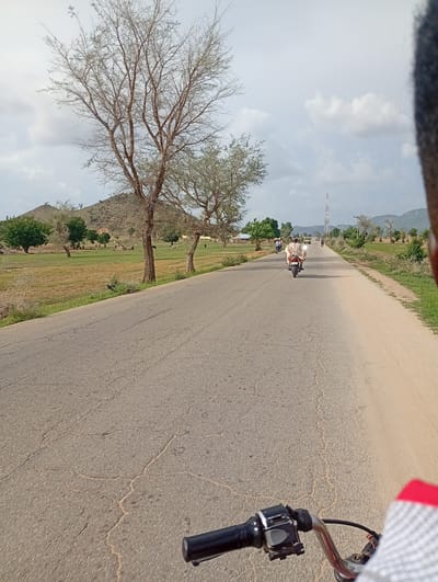 The image depicts a scene on a rural road in Shuwa, Nigeria. The perspective is from a vehicle, likely a motorcycle, with the handlebars visible in the foreground. The road is paved and shows signs of wear and tear, with cracks visible on the surface. The environment is characterized by dry, sparse vegetation and a few trees with bare branches. In the distance, a hill or mountain range can be seen, and there are some buildings or structures scattered across the landscape. Several people are present on the road, some riding motorcycles and others possibly walking. The sky is partly cloudy. The overall impression is of a dry, somewhat desolate rural area.