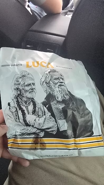 A white paper bag, branded "TRADIȚIE LUCA" in yellow and black text and featuring two black-and-white illustrations of elderly men with long hair and beards, is held in the lower-left by a human hand. Visible grease stains are present on the bag's lower section, across a design of three yellow horizontal stripes bordered by thinner black stripes. The website "www.lucatraditie.ro" is printed below these stripes. The scene is set within a vehicle in Costinești, Romania, indicated by dark grey textured car interior elements such as a center console or armrest and seat upholstery. A light-skinned limb is partially visible in the upper right background, and the lap of the individual holding the bag, clad in light brown fabric, is present in the lower right. No explicit interactions or additional individuals are discernible.