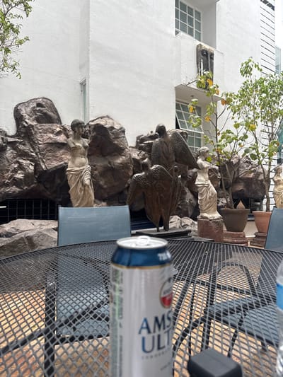 The image shows an outdoor scene with sculptures placed against a white building, possibly in Zapopan, Mexico. The sculptures appear to be of classical figures and eagles set amongst dark, rocky elements, simulating a natural environment. A small tree is also part of the arrangement. The foreground shows a metal mesh table, partially obscured by a can of Amstel Ultra beer. A blue chair with fabric seating is partially visible. The perspective is slightly tilted, suggesting the image was taken from a low angle.