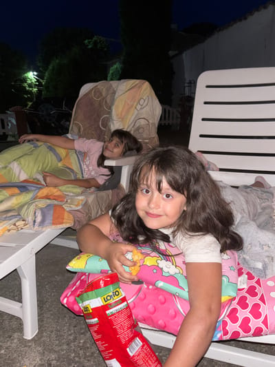 Two young girls are lying on a white lawn chair outdoors at night. The girl in the foreground, with long dark hair, is on her side and looking at the camera with a smile. She is wearing a white t-shirt with a pink design and is holding a snack. Next to her is a red bag of chips. The girl in the background is also lying on the chair, wearing a pink t-shirt. She is also looking towards the camera, with a slight smile. Both girls are covered with blankets. The setting appears to be a backyard with trees and lights visible in the background. The overall mood is relaxed and casual. There is visible text on the chip bag.