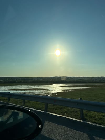 The image shows a view from a vehicle, possibly moving on a road. The sun is prominent in the sky, which transitions from light blue to a yellow-ish hue near the horizon. Alongside the road, there is a body of water reflecting the sunlight, potentially a flooded area or a lake, adjacent to some green land. A metal guardrail is visible next to the road. Part of the vehicle's side mirror is visible in the bottom right corner. The location is possibly in the area of Tarasovo, Russia, as indicated.
