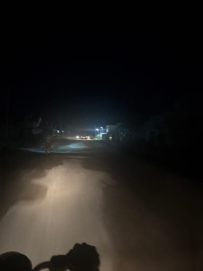 A nighttime scene captured from the vantage point of a moving vehicle, likely a motorcycle, traveling on an unpaved road in Angwan Sayawa, Nigeria. The lower portion of the image displays the dark silhouette of vehicle handlebars in the bottom-center and left. A powerful, warm-toned light source, consistent with a headlight, illuminates the immediate road surface, revealing light brown, dusty ground with visible patterns of disturbed particulate matter. The unpaved road extends into the midground, flanked by dimly visible, dark structures on both sides, presumably buildings. Multiple artificial light sources, including distant streetlights, are present further down the road, casting diffuse illumination. Two pairs of headlights from other vehicles are faintly visible in the distance. A noticeable atmospheric haze or dust diffuses the distant light sources. The upper two-thirds of the image consist of an undifferentiated, deep black expanse, indicating a dark night sky. No individuals are discernible.