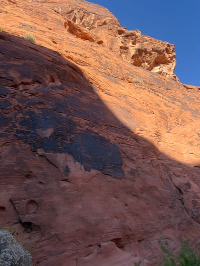 The image depicts a rock wall in the Valley of Fire State Park near Moapa Valley, Nevada, United States. The rock wall is primarily red and orange, with dark patches potentially caused by desert varnish. The left side of the wall is in direct sunlight, while a diagonal shadow bisects the wall. Petroglyphs are visible on the shaded portion of the rock face. To the left of the main wall is a rocky outcrop with similar coloring, silhouetted against a bright blue sky. Some vegetation is visible in the foreground and along the top right edge of the rock face.