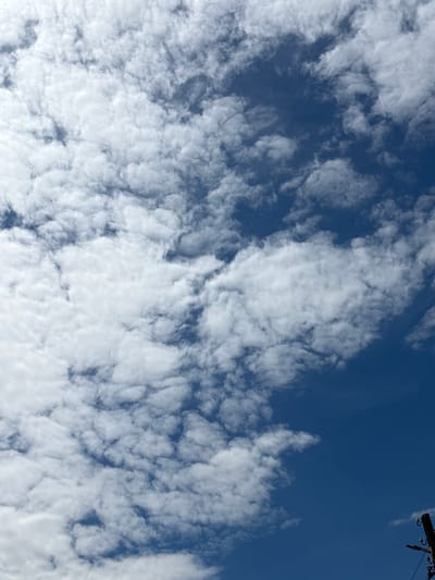 A clear, bright blue sky is dotted with fluffy, white cumulus clouds that stretch across the frame from the left side to the right. The clouds are scattered in an irregular pattern, with some areas appearing denser than others, creating variations in light and shadow. The bottom right corner of the image shows the top of a wooden utility pole, with insulators and wires visible, suggesting an outdoor urban or suburban setting. The image captures a typical daytime sky, with no people or discernible human activity beyond the presence of the utility pole.