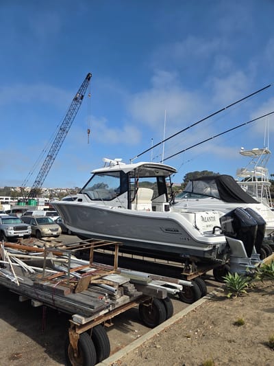 The image depicts a boatyard in Dana Point, California, USA. In the foreground, a flatbed trailer is laden with various construction materials, including lumber, pipes, and metal framing. To the right, two agave plants are visible. Behind the trailer, a white boat with a gray hull is prominently displayed on a trailer. The boat has two black Mercury outboard motors attached to its stern and the name "PALANTIR" is visible on the transom. Further back, another white boat with a t-top is partially obscured. A large industrial crane with a dark blue lattice structure is positioned in the background, extending into the bright blue sky with scattered clouds. Several vehicles, including silver SUVs and a minivan, are parked behind the boats. Residential buildings with red-tiled roofs are visible on a distant hillside. The ground is covered with gravel and dirt.