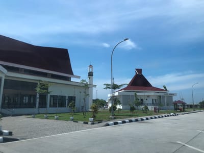 The image shows a street view of a building complex with two main structures, likely a mosque and an accompanying building, under a clear blue sky with scattered clouds. The larger building on the left has a long, angled, dark red roof and a white facade with rows of windows. In front of this building, a paved area transitions to a grassy area with several potted trees. A tall, slender minaret with a white exterior and a red-capped roof stands between the two main buildings. The building on the right is smaller, featuring a prominent, steeply pitched dark red roof and a white facade with decorative grilles and windows. A small gazebo with a red roof is visible to the right of this structure. A street lamp with a curved pole stands between the buildings. A paved road with black and white striped curbing runs across the foreground. Two individuals, one in the distance near the larger building and another closer to the right-hand building, are visible. The scene is set in an open area with some vegetation and paved surfaces, suggesting an institutional or public space in Gohor Lama, Indonesia.