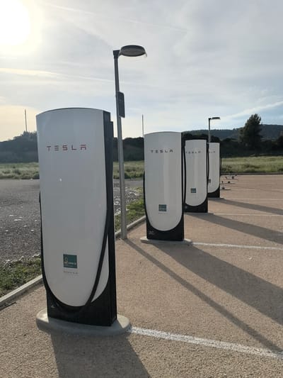 The image displays an outdoor electric vehicle charging station featuring a row of four Tesla Supercharger stalls under a bright, low-angled sun. The time of day appears to be either early morning or late afternoon, as indicated by the long, distinct shadows cast by the chargers and lampposts across the ground.
The foreground is dominated by the first Tesla Supercharger, a tall, sleek unit with a white body and a black base/trim. The word "TESLA" is prominently displayed in red capital letters on its upper front. Below this, a green square logo with the text "moves III" and "energía y movilidad" is visible. Near the bottom, the designation "2C" is marked. A modern lamppost stands immediately next to this charger, and another lamppost can be seen further down the line of chargers.
The ground is a paved surface, likely asphalt or gravelly asphalt, with white lines marking parking spaces. In the mid-ground, three more identical Tesla Superchargers recede into the distance, each bearing the "TESLA" logo. Beyond the charging area, the background features a natural landscape of green scrubland, grass, and rolling hills covered with trees. The sky above is bright and clear, with a few faint wisps of clouds or contrails. There are no people or vehicles visible in the scene, conveying a sense of quiet.
