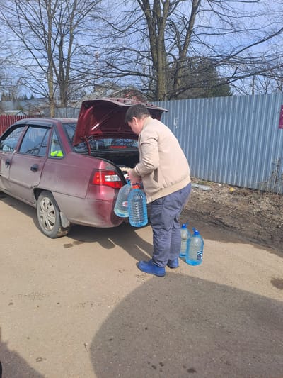 In Avdot’ino, Russia, a daytime scene depicts a male subject loading or handling large clear plastic water bottles near the open trunk of a maroon four-door sedan. The individual, dressed in a tan jacket, dark blue trousers, and blue shoes, is seen holding two large, approximately five-gallon, bottles filled with a light blue liquid, while two identical bottles rest on the unpaved road surface beside him. The sedan, consistent with a Daewoo Nexia model, shows visible dirt on its exterior. The environment consists of a dirt and gravel road with scattered muddy patches, particularly near a light grey corrugated metal fence that runs parallel to the road in the mid-ground. Beyond the fence, a landscape of bare deciduous trees stands against a light blue sky, suggesting a non-summer season. A segment of a red wooden fence is visible on the far left, and a small red sign is affixed to the corrugated fence on the right.