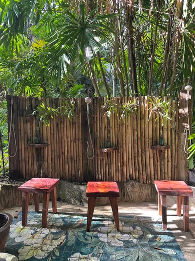 The image depicts an outdoor shower area with a bamboo wall in Khlong Thom Nuea, Thailand. Three wooden stools with reddish tops are placed on a tiled floor with a floral pattern. Behind the stools, the bamboo wall has three showerheads with hoses and small wooden shelves, each holding a green bottle. Plants are growing from the bamboo wall. The background consists of lush green foliage and trees. The scene is illuminated by sunlight, creating shadows on the ground.