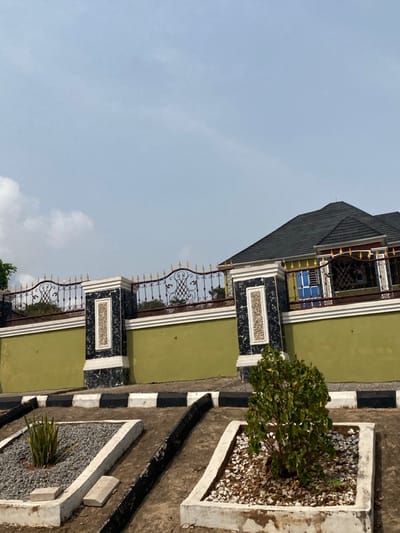 This image captures an exterior scene in Ebuwana, Nigeria, under a bright, partly cloudy sky. The prominent feature is a well-maintained compound wall in the midground, constructed with an olive-green lower section, topped by a white decorative band. Multiple large pillars clad in dark, marble-patterned material with white veining are integrated into the wall, each containing a recessed rectangular panel with a textured, light-colored infill. Above the solid wall and pillars, an ornate wrought iron fence with black scrollwork and numerous golden finials extends across the frame. Behind this fence on the right, a building with a dark grey, multi-pitched roof is partially visible, featuring a prominent window with a blue frame. The foreground consists of dry, light brown earth, two raised rectangular garden beds made of white concrete. The left bed contains grey gravel and a single Sansevieria plant, accompanied by two small concrete blocks. The right bed holds a small, leafy green bush surrounded by a mix of white stones and brown mulch. Black-and-white striped curbs separate the garden beds from the compound wall, and dark-colored drainage channels run along the ground. No individuals are present in the scene.
