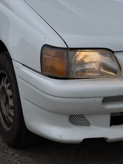 The image displays a close-up, angled view of the front right side of a stationary white passenger vehicle in Mogadouro, Portugal. The vehicle's right headlight is illuminated, emitting a warm, yellowish light through a ribbed, clear plastic lens. Regulatory markings, including "E6," "S437," and "HCR," are visible on the headlight lens. To the immediate left of the main headlight, an amber turn signal lens is integrated into the assembly. The white front bumper shows general dirt accumulation and exhibits minor green paint transfer or scratching on its lower left section. A black, perforated mesh grille is recessed into the lower right portion of the bumper. The white front fender and a portion of the hood are visible, with some dirt and a small dark speck present on the fender above the headlight. The front left wheel, featuring a dirty, silver-colored, multi-spoke rim, and its black tire are partially visible on the far left. The vehicle is parked on a dark, coarse gravel surface. No people or interactions are visible.