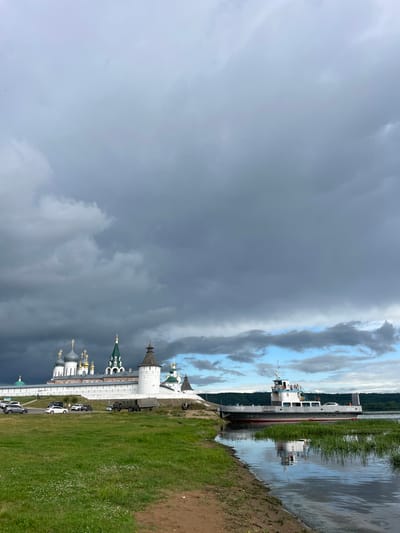 This image depicts the Trinity Monastery of Saint Macarius in Makar'evo, Russia, situated on the banks of a wide river. The monastery complex is characterized by its white stone walls, multiple domed churches with gold and green spires, and defensive towers. In the foreground, a ferryboat with the designation "СП-35" is docked near the grassy bank. The river reflects the boat and the sky, which is filled with dramatic, dark, and light grey clouds, suggesting an overcast or approaching storm. On the bank near the monastery, several vehicles are parked, including cars and a military-style truck. A few individuals are visible near the vehicles and along the monastery walls. The landscape includes green grass and a sandy shoreline.