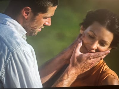 In this close-up image, a man with short, dark hair and a light-colored collared shirt is holding the face of a woman with short, dark, curly hair. Both individuals appear to have injuries, with visible cuts and abrasions on their faces and the man's hands. The man's right hand is cupping the woman's chin and cheek, while his left arm is resting on her shoulder. The woman's eyes are closed, and her head is tilted downwards, suggesting distress or pain. The background is a blurry, green, outdoor environment. The image does not provide any information about the city of Высокая Гора, Russia, or any context related to this location.