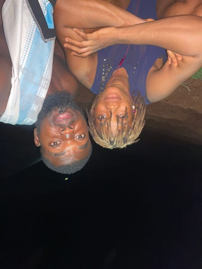 Two people pose in an outdoor nighttime scene. The man on top has dark hair, a beard, and wears a light blue and white patterned shirt. Below him, a person with blonde dreadlocks wears a blue shirt and pink earbuds. The location appears to be in Ogbe, Nigeria, as indicated by the provided context. The background is dark, possibly a field or park.