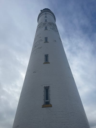 The image displays a tall, conical lighthouse situated in Montrose, United Kingdom. The structure is constructed from brick and painted white, exhibiting noticeable weathering with areas of peeling paint and exposed darker brickwork, particularly on its upper sections. Five vertically elongated, rectangular windows with dark frames and prominent yellow sills are evenly spaced along the tower's visible length. The lighthouse tapers upwards from a broader base to a narrower point, where a dark, presumably metal, gallery or railing system is visible just below the unpictured lantern room. The photograph is taken from a low-angle perspective, emphasizing the lighthouse's height against a partially overcast sky featuring a mixture of grey clouds and patches of light blue. No individuals, vehicles, or other ground-level objects are visible within the frame.