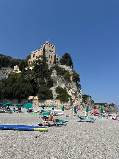 The image depicts a sunny day at a pebble beach in Finale Ligure, Italy, with a prominent castle-like structure perched atop a cliff in the background. In the foreground, a blue and yellow stand-up paddleboard lies on the beach. Several lounge chairs and umbrellas are set up on the pebble shore, with a few people sunbathing or relaxing. The castle, appearing to be a historic building with a crenellated roofline, overlooks the beach and the Mediterranean Sea. Lush green trees and rocky terrain surround the castle. The sky is a clear, bright blue.
