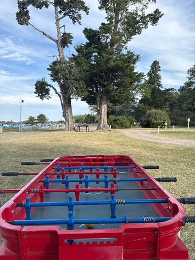 A foosball table stands in a park-like setting. The table is red with blue and red players, and black handles. Behind it, a grassy area extends towards large trees, with a clear blue sky overhead. In the distance, benches and people can be seen, along with a row of lights. The scene conveys a casual, outdoor recreational area.