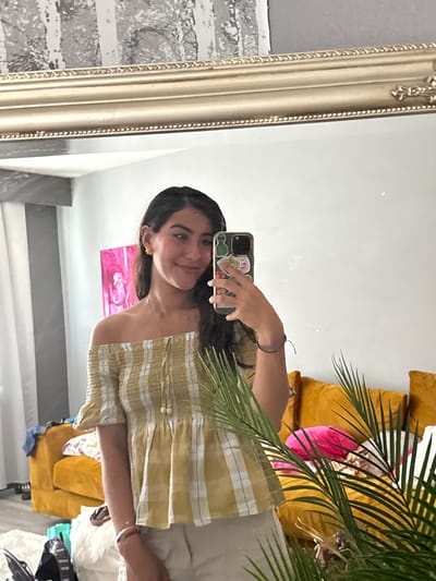 A young woman is taking a mirror selfie in a room. She is wearing a yellow and white striped off-the-shoulder top. Her dark hair is long and down, and she is smiling at her phone. In the background, a large gold-framed mirror reflects the room. A vibrant yellow couch with decorative pillows and a pink accent pillow is visible in the reflection. A large potted plant with green fronds is in the foreground, partially obscuring the woman and the reflection. The room appears to be well-lit, suggesting daytime. The style of the room and decor suggests a home environment.