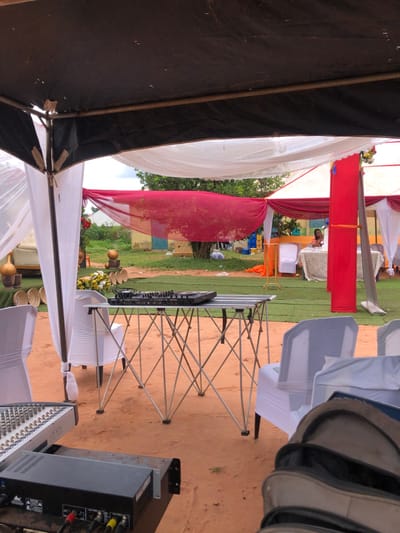 The image displays an outdoor event setup in Amanuke, Nigeria. The scene is illuminated by natural light, indicating daytime. The ground consists of reddish-brown soil with interspersed sections of artificial green turf.
In the immediate foreground on the left, a black audio amplifier with visible red, yellow, and white RCA cable connections is present. To the right, a beige bag is partially visible.
Centrally positioned in the mid-ground is a silver-colored, collapsible folding table, on top of which rests a black DJ controller equipped with numerous knobs, faders, and buttons. White chairs, many covered with white fabric slipcovers, are arranged throughout the foreground and mid-ground. Overhead, black and white fabric panels form part of a canopy structure, supported by metal poles. Additional decorative elements include drapes of red and white sheer fabric stretched across the scene.
To the left in the background, a decorated area features several brown gourds or calabashes, some placed on stands, and multiple flat, circular woven baskets. A white upholstered armchair and white floral arrangements are also visible here. To the right, a single individual, appearing to be a light-skinned person with dark hair and wearing an orange or peach-colored top, is seated at a table covered with a white tablecloth. Another white chair is positioned adjacent to this table.
Further in the distant background, a yellow-painted building with a blue door is discernible, surrounded by green trees and foliage. A stack of blue crates is visible near the building.