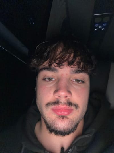 A young man with olive skin, light brown eyes, and dark, curly hair is facing the camera. He has a beard and mustache. His lips are full and appear slightly glossy. He is wearing a dark-colored hoodie or jacket. The lighting is dim, and there are some out-of-focus objects in the background. The image appears to be taken indoors, possibly in a car or dimly lit room.