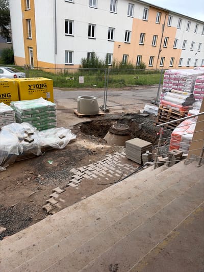 The image depicts a construction site in the city of Příbram, Czechia, adjacent to a residential building. The building, painted in white and orange, has multiple windows. In the foreground, concrete stairs lead up from a construction area filled with rubble, gravel, and building materials. A deep excavation is visible, revealing a manhole cover. Several stacks of building supplies, including bags of mortar and insulation materials, are present. A concrete ring and pallets of building materials are also visible. The construction site is enclosed by a metal fence, with grass and trees in the background.