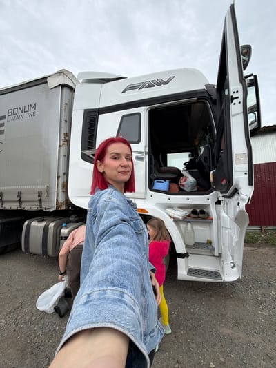 This portrait-oriented photograph captures a woman and a child beside a large white semi-truck and trailer under an overcast sky. The scene is set outdoors on a gravel surface, with a reddish-brown metal-clad building visible in the background.
In the foreground, a woman with short, bright red hair, wearing a denim jacket over a light-colored top, poses for the camera with a subtle smile. She appears to be holding the camera for a selfie or taking the photo from a low angle. Her left arm is visible in the bottom right corner of the frame.
To her right, partially obscured by the truck and her body, a child in a pink long-sleeved top and bright yellow leggings is seen crouching or kneeling near the truck's side. The child's upper body, legs, and feet in light-colored sneakers are visible, and a white plastic bag rests on the ground nearby.
The central subject is a white FAW brand semi-truck. Its driver's side door is open, revealing the cab's interior, which contains various items including bags and a blue container. External storage compartments near the front wheel are also open, housing items like shoes, a white plastic bottle, and what appears to be another plastic bag. The truck is connected to a silver-colored trailer with "BONUM MAIN LINE" written on its side. The trailer's upper frame shows visible rust.
The image suggests a stop or break during a journey, possibly at a truck stop or yard in Kamyennoye, Russia. The overcast sky indicates daytime without direct sun.