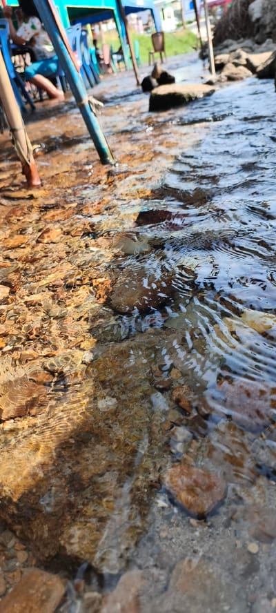 A low-angle, forensic perspective captures a shallow, clear stream in Aeknatolijulu, Indonesia, with water flowing over a bed of multicolored rocks and pebbles. Sunlight refracts through the water, creating ripples and illuminating the streambed. Two vertical support poles are partially submerged in the mid-ground: one on the left is natural wood-colored with visible bindings, and the other on the right is painted blue. In the blurred background, under a blue canopy structure, a person is seated with their lower legs and feet submerged in the water; they wear light blue shorts and a light-colored, patterned top. Several blue plastic chairs are visible near the seated individual. Further to the right in the background, a single brown chair rests on a patch of green grass, adjacent to a large pile of dry organic material.