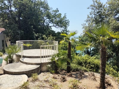 The image depicts an outdoor scene featuring a circular, multi-tiered concrete patio with a white railing. The patio is surrounded by lush greenery, including several palm trees of varying sizes and other dense foliage. In the background, glimpses of the sea can be seen through the trees, suggesting a coastal location. The sky is clear and blue, indicating fair weather and likely daytime.

To the left of the patio, a portion of a building with a tiled roof is visible, along with a streetlamp. The ground around the patio is covered in gravel and small plants. The overall setting appears to be a garden or recreational area, possibly associated with a residence or a public space.

A timestamp in the bottom left corner reads "2026.06.29 13:19," indicating the date and time the photograph was taken. The image conveys a sense of tranquility and natural beauty, enhanced by the bright sunlight and the vibrant green vegetation. The location context of Krk, Croatia, aligns with the presence of palm trees and proximity to the sea. There are no people visible in the frame, focusing the attention on the architectural element and the natural environment.