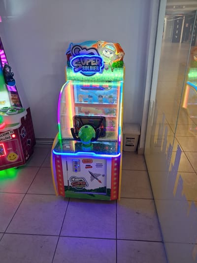 A brightly lit, vertically oriented arcade game cabinet with a "SUPER SOLDIER" theme stands in an indoor public space, likely a mall or amusement center, as suggested by the tiled floor and reflective surfaces. The game has a green joystick and a ticket dispenser. To the left, another arcade game cabinet is partially visible, with a sign that reads "WELCOME TO CHALLENGE." A white trash receptacle is located to the right of the "SUPER SOLDIER" game, against a plain wall. The lighting is a mix of natural and artificial, with colorful LED lights illuminating the game cabinets. The overall environment appears clean and well-maintained. There are no people visible in the image. This scene is consistent with a commercial entertainment area in Constanța, Romania, given the typical setup of such venues in the city.