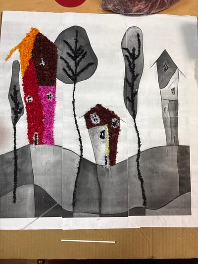 This is a collage artwork depicting a whimsical village scene, likely created by children. The artwork features several stylized houses made of colorful shredded paper, including houses in bright red, pink, and orange. These houses are interspersed with black, twig-like trees and set against a background of grayscale abstract shapes that suggest rolling hills or landforms. Some of the houses have small, dark windows. Inscriptions in what appears to be Cyrillic script are present on some of the grayscale elements, possibly labeling colors or elements of the scene. The artwork is mounted on paper and appears to be a composite of several sheets. In the foreground, a portion of a cardboard box is visible, and two white sticks, possibly straws or similar items, are laid across the bottom of the artwork. There are no people or animals visible in the image. The artwork is likely located in a home or classroom setting in Ветрынскі сельскі Савет, Belarus, given the presence of Cyrillic script and common artistic materials.