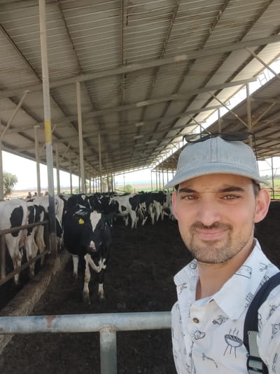 In Yoav Regional Council, Israel, a man wearing a light blue denim cap and a white patterned shirt is taking a selfie in a cow farm. The man has short brown hair and a light beard. He is smiling and looking at the camera. Behind him, a herd of black and white cows are standing in a muddy enclosure, under a large metal roof supported by poles. The scene suggests a warm, sunny day.