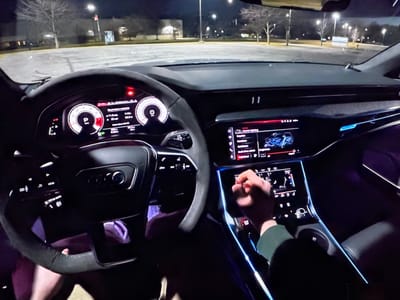 The image shows the interior of a car at night, likely in a parking lot, with the dashboard and steering wheel prominently displayed. The car's digital dashboard shows speed and other vehicle information, and the central infotainment screen is also visible, displaying interior lighting options. A person's hand is shown interacting with the touchscreen. The background reveals a dark, paved area with trees and streetlights, suggesting an urban or suburban setting. There are no identifiable landmarks that would place the image specifically in Bezden, Bulgaria. The scene is static and provides no information about ongoing events or specific activities beyond the interaction with the car's controls.