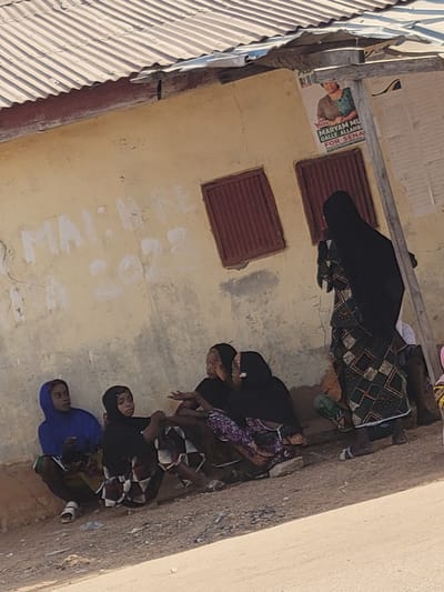 This image depicts a street scene in Nassarawa Egon, Nigeria, featuring six individuals gathered by a weathered building. The building wall is a light yellow-ochre color, exhibiting cracks and discoloration, with the text "MALIHA" and "A 2022" spray-painted in white. Above, a corrugated metal roof is visible, with its underside painted dark red. Two reddish-brown, possibly louvered, windows are set into the wall. A wooden utility pole stands on the right, affixed to which are political posters, one prominently displaying "Vote MARYAM MU GALLE ALLAHB FOR SENA."

On the unpaved, dirt ground bordering a light-colored road or curb, a group of five individuals, primarily females, are seated or crouching. From left to right: a young female in a blue hijab and patterned skirt sits with white sandals. Next to her, a younger female child in a black hijab and a dark, checkered dress also crouches. Two adult females are seated closely together; both wear dark hijabs, with one in a purple-patterned garment and the other in a darker patterned dress. They appear to be engaged in a discussion, with one gesturing with her hand. Partially obscured behind this group is a fifth individual.

To the right of the seated group, a sixth individual, an adult female, stands facing towards the left. She wears a long black hijab that covers her head and body, over a distinct patterned dress featuring green, black, and beige geometric (square and diamond) shapes. She is wearing light-colored sandals. The ground beneath the individuals contains small pieces of visible detritus. A person with a pink head covering is partially visible at the far right edge of the frame.