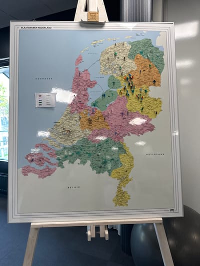 The image shows a large map of the Netherlands titled "PLAATSNAME NEDERLAND", mounted on an easel. The map is divided into provinces, each with a different color and a two-letter abbreviation. Numerous small pins in various colors are stuck into different locations on the map, indicating specific places. A legend is visible in the lower left corner, with color-coded entries next to names, suggesting the pins represent individuals or categories. The map itself is detailed, showing cities and towns. The background includes a portion of a building's exterior with windows on the left and a wall on the right, suggesting an indoor setting. A black exercise ball is visible in the lower right corner. The city of Staphorst, Netherlands is not explicitly labeled on this map, nor is it visually identifiable as a specific location within the frame.