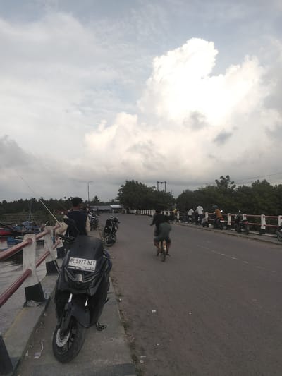 In Tambonbaroh, Indonesia, an overcast sky with large, white cumulus clouds hangs over a scene featuring a bridge, a road, and a body of water. Several people are present. On the bridge to the left, a person stands near fishing rods, looking towards the water where several boats are moored. Nearby, a black scooter is parked.
Further along the bridge and road, multiple people are seen. Some are leaning on the bridge railing, while others are interacting or standing. A person rides a bicycle on the road, and several motorcycles are parked on the side of the road. The road itself appears to be an asphalt surface with lane markings. Trees line the opposite side of the road, forming a backdrop to the scene. The overall impression is of a moderately populated area with a mix of recreational and transport activities.