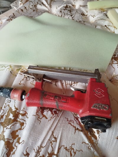 The image shows a close-up view of a work-in-progress scene, likely related to upholstery or crafting.
The most prominent object is a well-used, red pneumatic stapler, positioned diagonally across the frame. It features a metallic silver magazine for staples along its top, and a black trigger. The stapler appears to be air-powered, with a black and red air hose fitting attached to its rear. The red body of the stapler shows signs of wear, with scuffs, scratches, and some darker, possibly greasy, smudges. On the side of the stapler, faint white text or a logo is visible, which looks like "CHKD" or similar block letters, and another symbol further back that resembles a stylized "S" or "SG" within a box.
Beneath and behind the stapler is a large, irregularly shaped piece of light green foam. It has a smooth, slightly textured surface and appears somewhat thick and pliable. In the upper right corner of the image, two smaller, rectangular pieces of light yellow foam are visible, suggesting they might be cut-offs or additional material.
The entire scene rests on a cream or light beige fabric with a distinctive golden, metallic, or glossy pattern. The pattern is abstract and flowing, resembling swirling branches, veins, or organic shapes that spread across the fabric. The fabric itself shows some wrinkles and folds, indicating it's draped or gathered.
The overall impression is one of a DIY or professional crafting project, specifically involving the use of foam padding and fabric, with the stapler being a key tool for assembly. The lighting suggests an indoor setting.