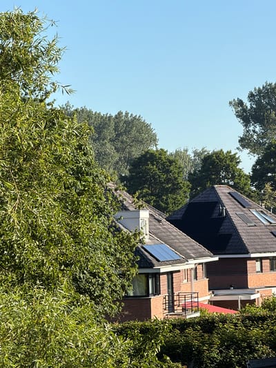 The image depicts a view of residential buildings in Druten, Netherlands, with lush green foliage in the foreground and midground, and a clear blue sky overhead. Two houses are visible, both with tiled roofs and brick exteriors. The house on the right has a dark, multi-faceted roof adorned with solar panels, and a prominent dormer window. The house on the left, partially obscured by a large, leafy tree on the left side of the frame, also features a tiled roof with solar panels. A red awning or canopy is visible extending from the lower part of the right house. The environment suggests a sunny day, possibly in the late morning or early afternoon, with the vegetation appearing healthy and vibrant. No people or animals are present in the image. The architectural style suggests a modern suburban setting.