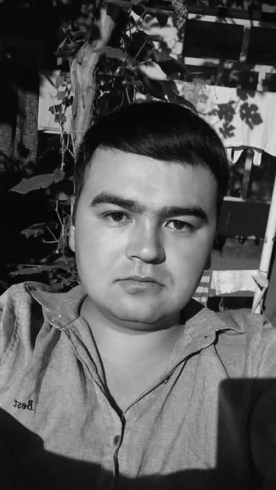 The image is a black and white, front-facing selfie of a young man with dark hair and a fair complexion. He is wearing a light-colored, collared shirt with buttons. His facial expression is neutral. The background appears to be outdoors, with foliage and what looks like laundry hanging on a line. There is a shadow cast across the man's shirt. The image is taken in the city of Kasansay, Uzbekistan.